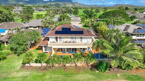 Una casa grande con piscina, rodeada de exuberante follaje y palmeras, con paneles solares en el techo.