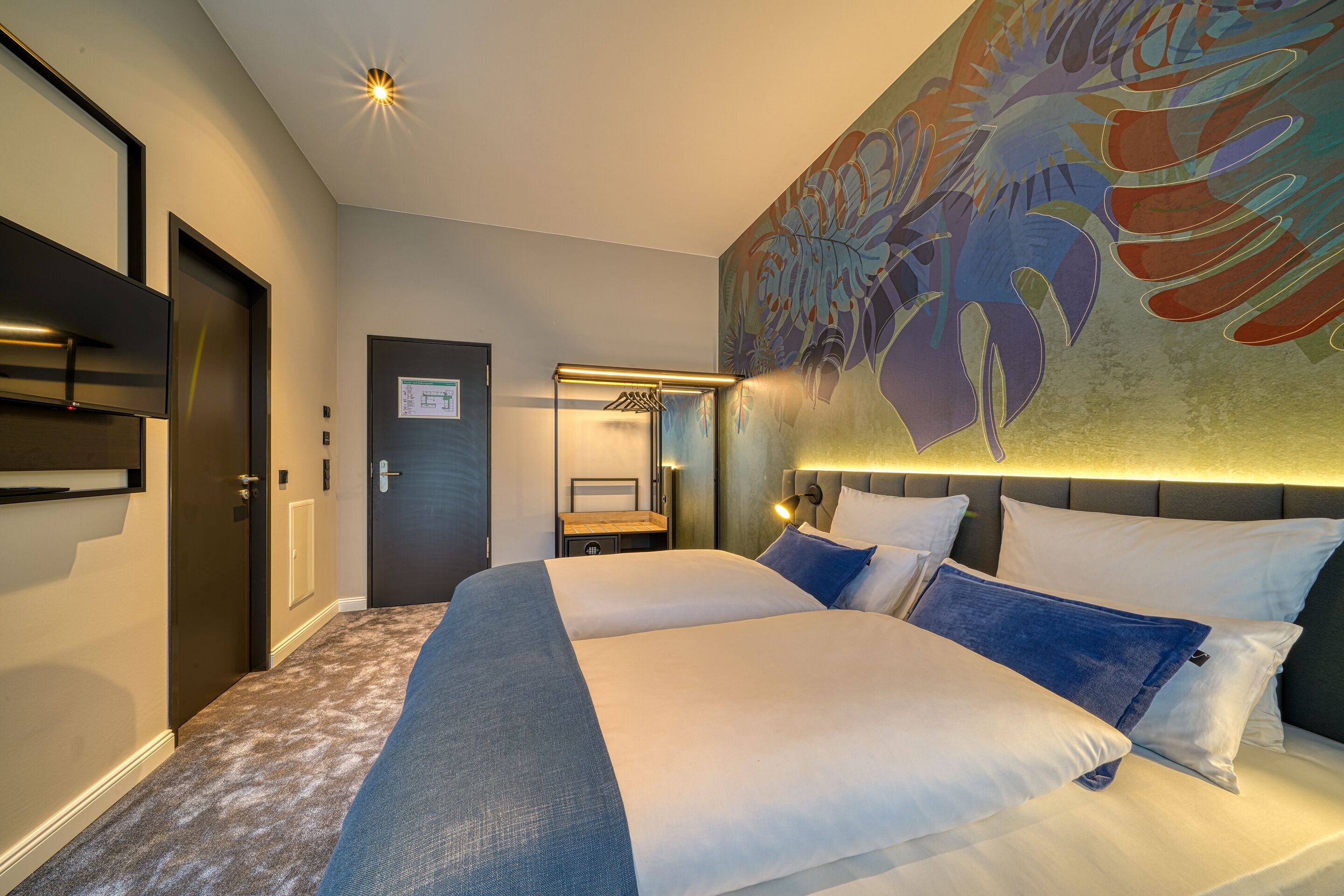 Una habitación de hotel con una cama grande, un cabecero con lámpara y un televisor montado en la pared, además de una puerta con un letrero.