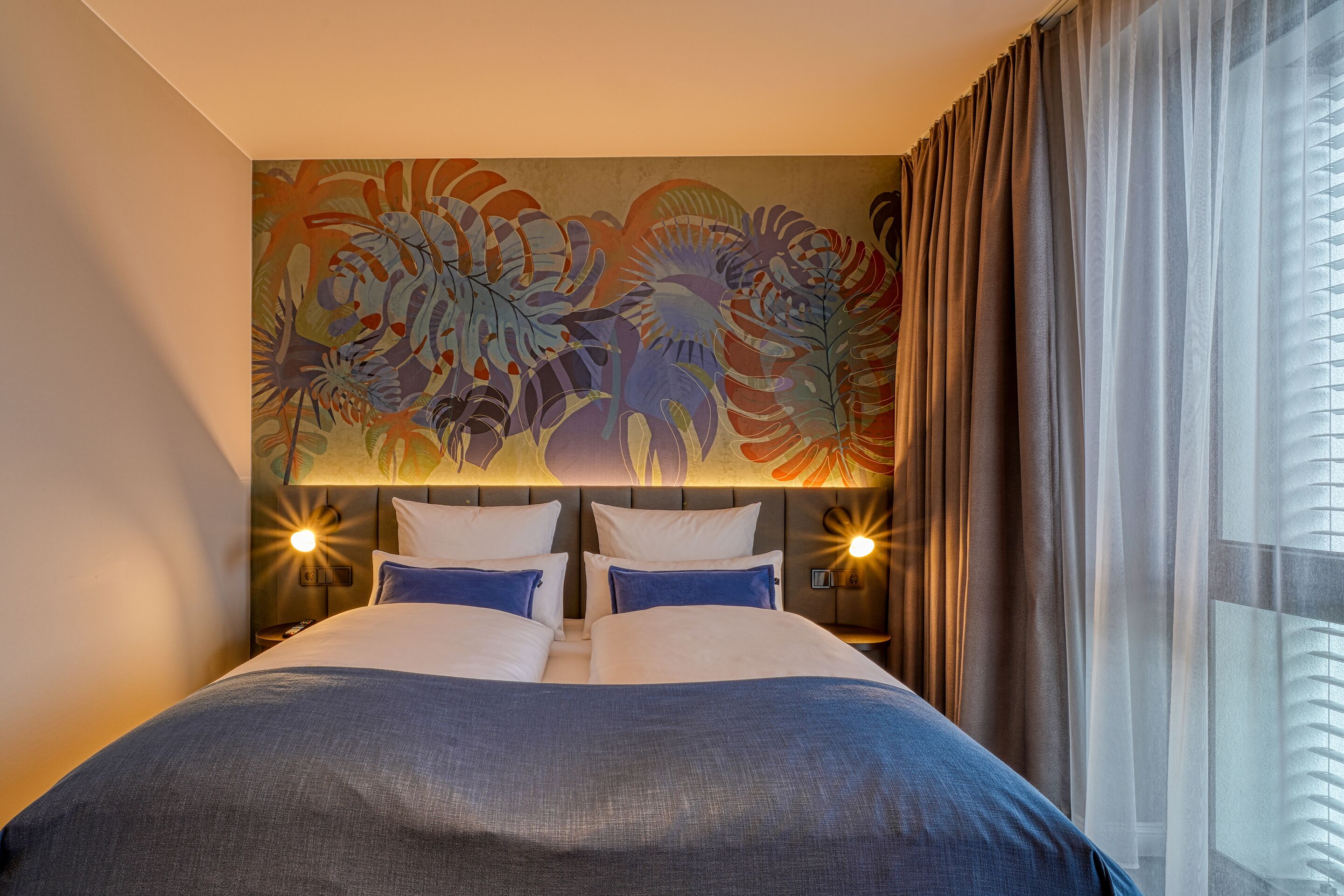 A hotel room with a large bed, two bedside tables, a headboard with a floral mural, and a window with curtains.
