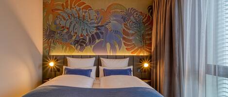 Une chambre d’hôtel avec un grand lit, deux tables de chevet, une tête de lit ornée d’une fresque florale et une fenêtre avec des rideaux.