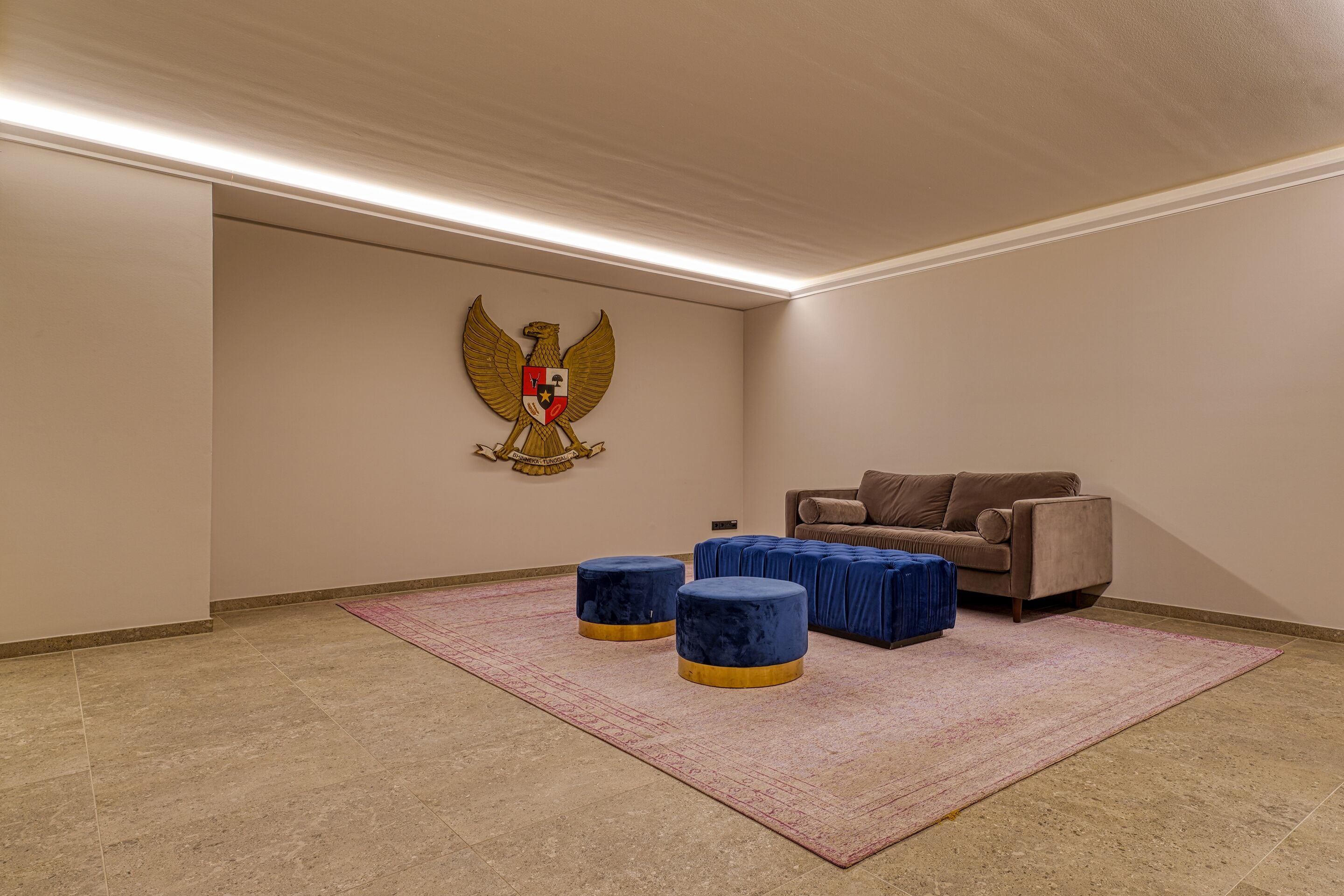 A room with a beige wall, a large emblem, a brown sofa, and two blue ottomans on a pink rug.