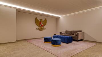 Una habitación con pared color beige, un gran emblema, un sofá marrón y dos otomanas azules sobre una alfombra rosa.
