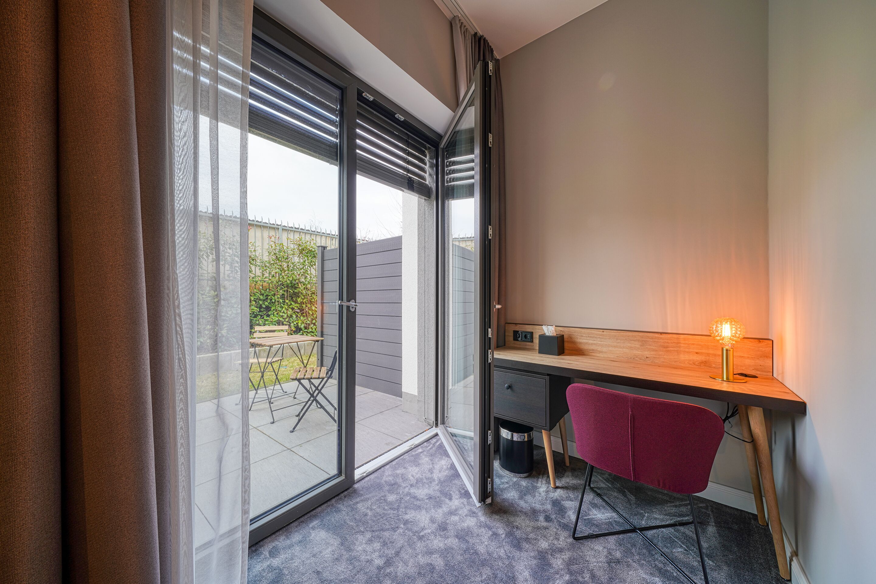 Une chambre moderne dotée d’une porte-fenêtre coulissante donnant sur un espace extérieur, d’un bureau en bois avec une chaise et d’une petite table avec une lampe.