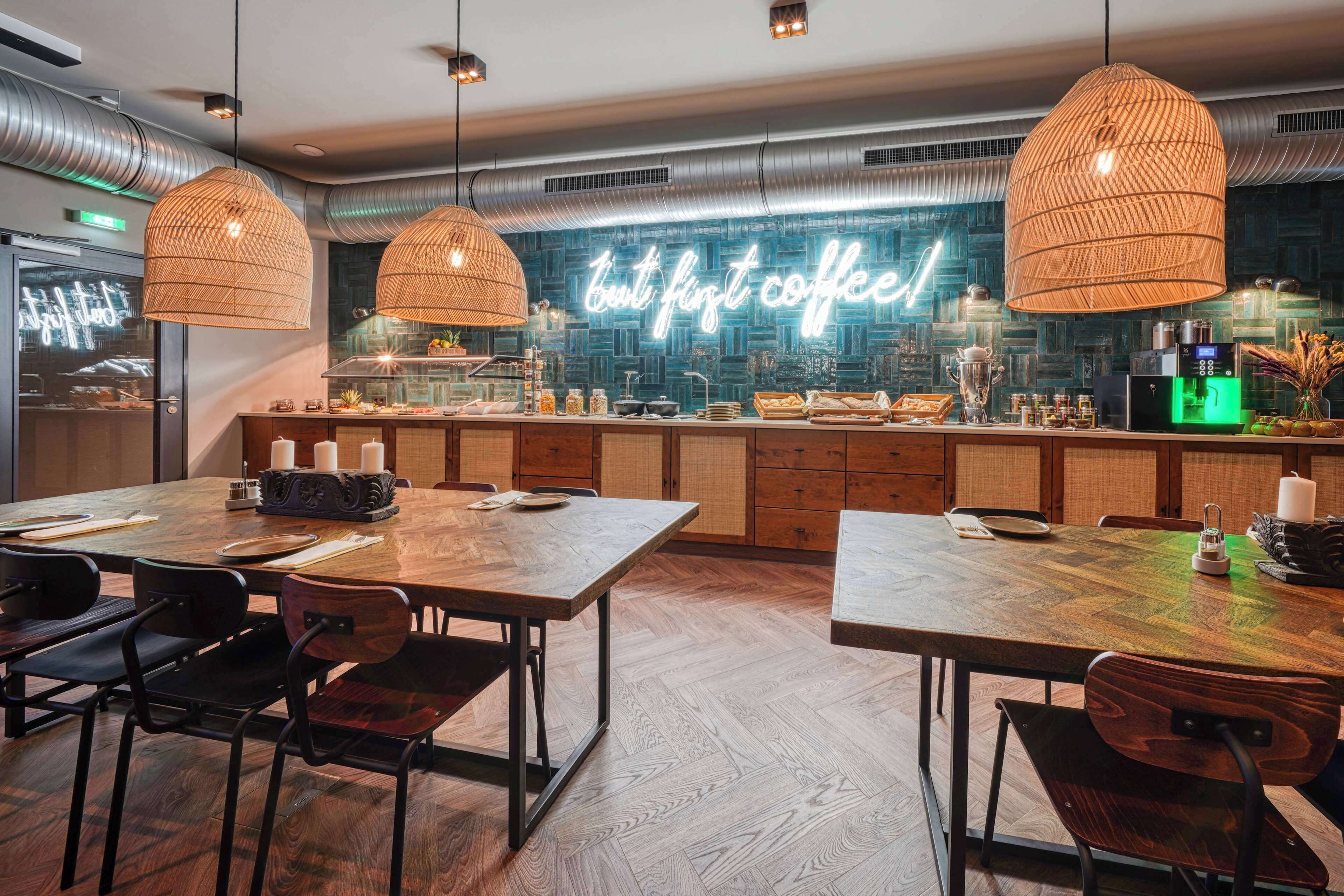 Ein modernes Café mit Holztischen und -stühlen, einem Neon-Schild mit der Aufschrift "first coffee" und einer Theke mit verschiedenen Kaffeemaschinen.