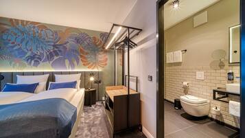 A modern hotel room with a large bed, a bedside table, a nightstand, a wooden bench, a wall-mounted TV, a bathroom with a toilet, sink, mirror, and towel rack.
