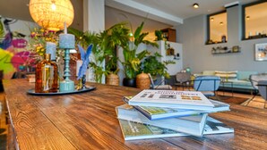 Una mesa de madera con libros y objetos decorativos, rodeada de plantas en macetas y una lámpara colgante.