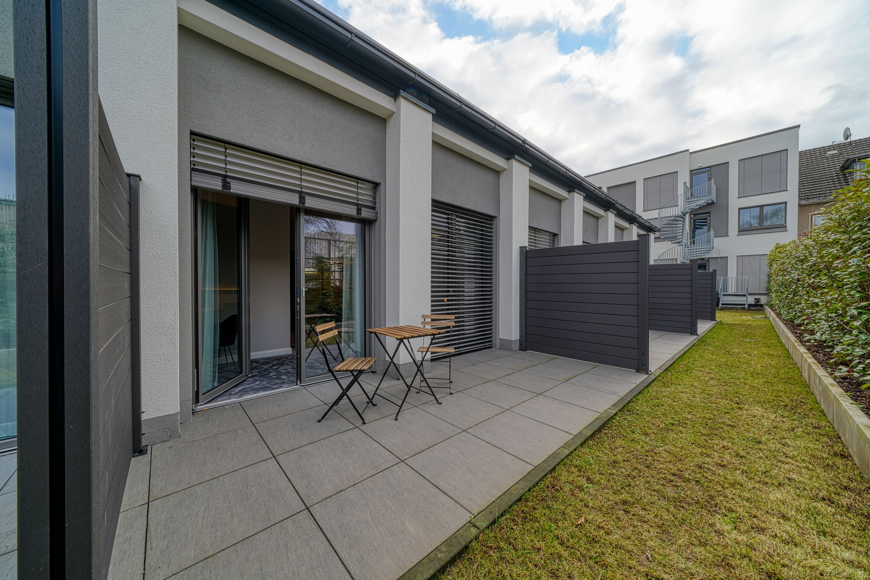 A modern residential building with a patio area featuring a table and chairs, a sliding glass door, and a small garden.