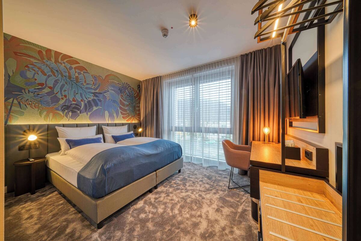 Quarto de hotel moderno com uma cama grande, uma escrivaninha com cadeira, televisão e uma janela ampla com cortinas.