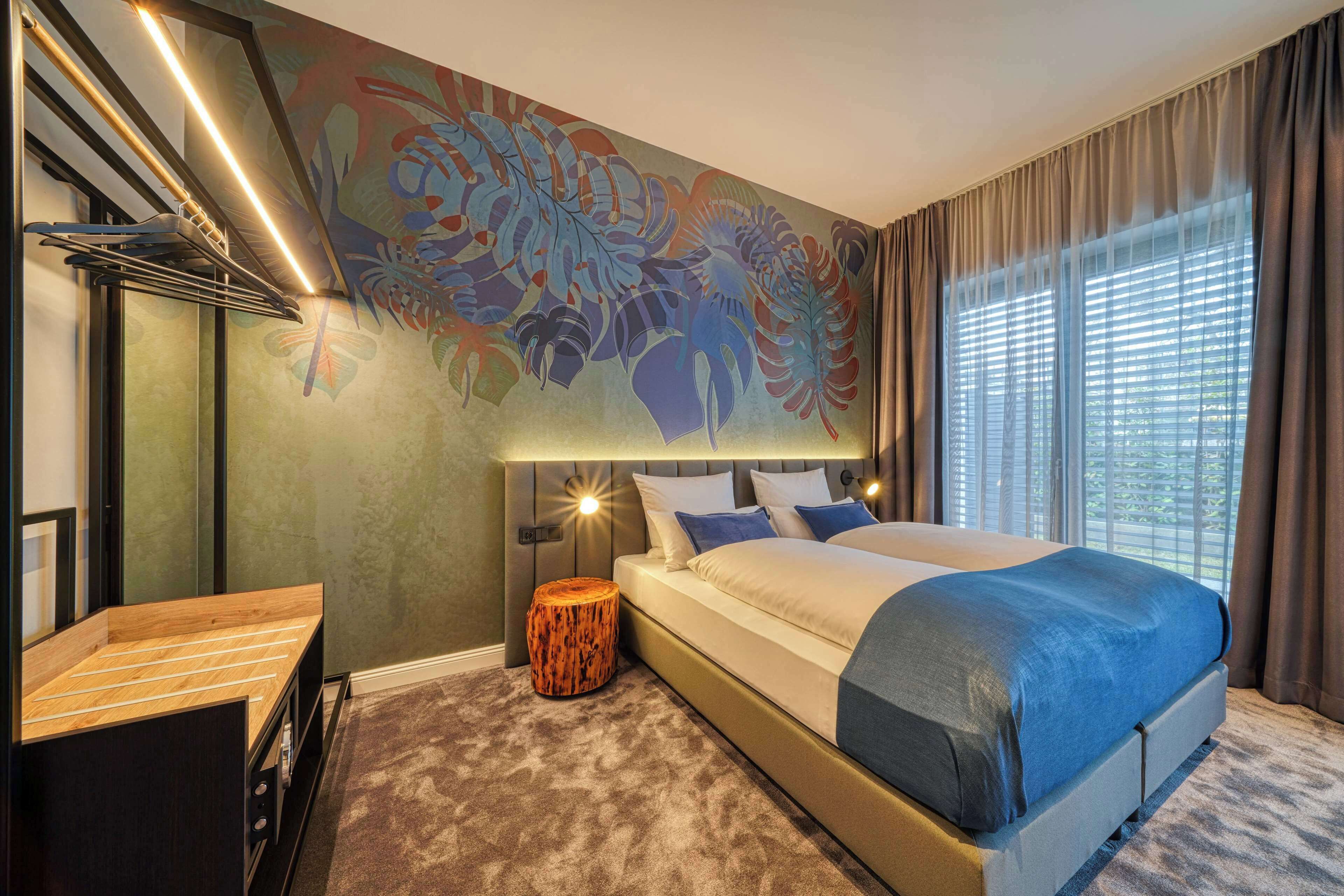 A modern hotel room with a large bed, a wooden side table, a round wooden stool, a wall mural, and a large window with curtains.