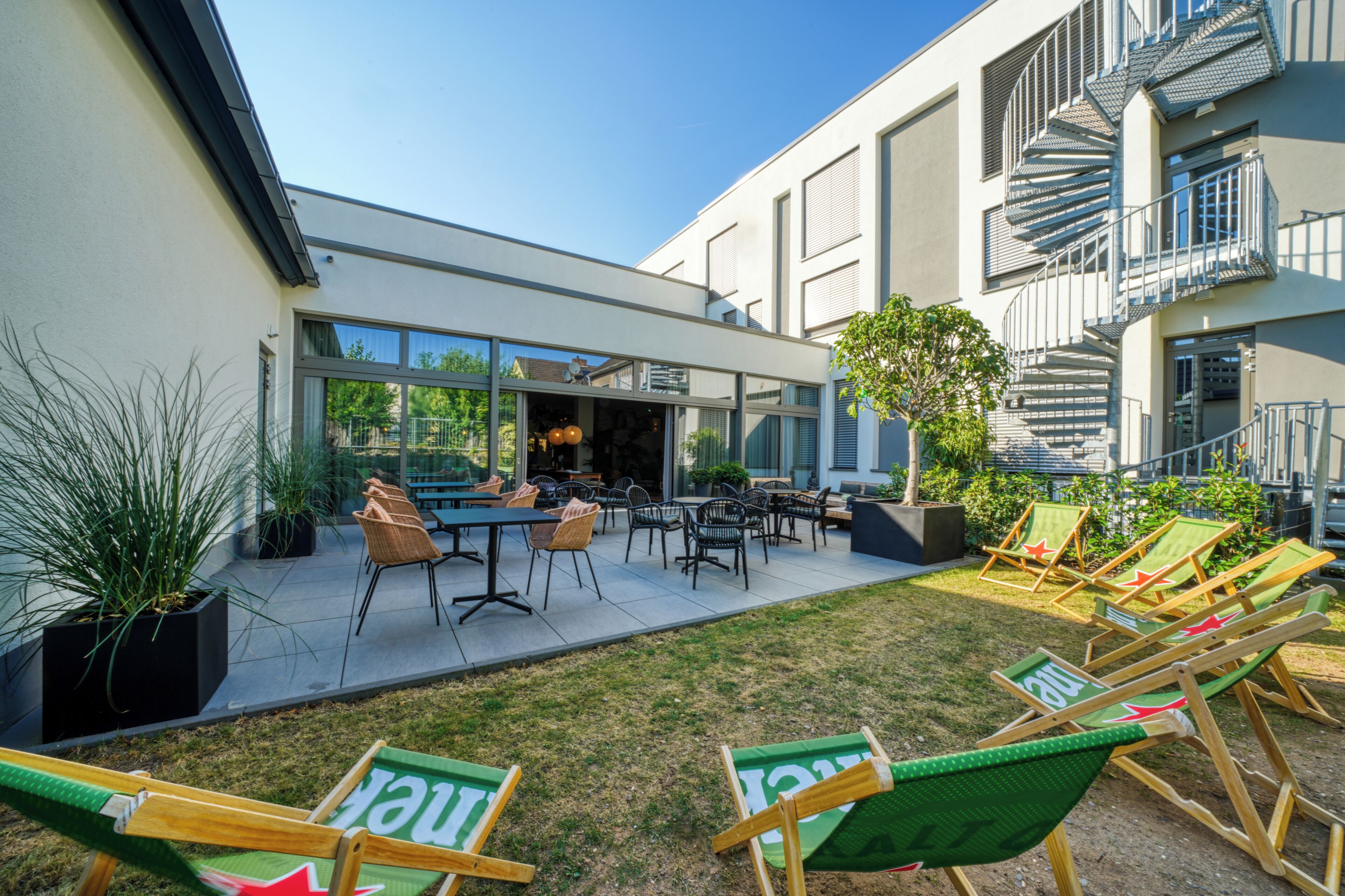 Un espace extérieur moderne aménagé avec des tables et des chaises, une pelouse verdoyante et un bâtiment doté de grandes fenêtres et d’un escalier.