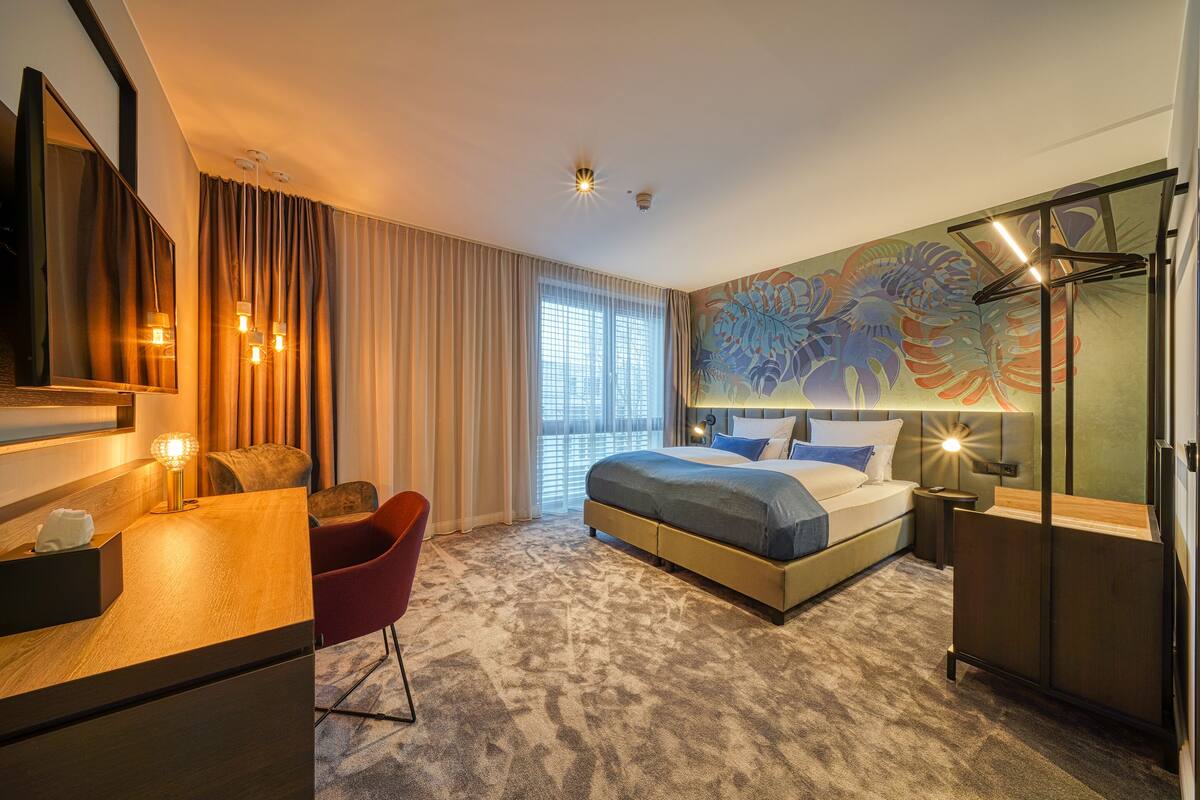 Quarto de hotel moderno com uma cama grande, uma escrivaninha, uma cadeira vermelha e uma janela ampla com cortinas.