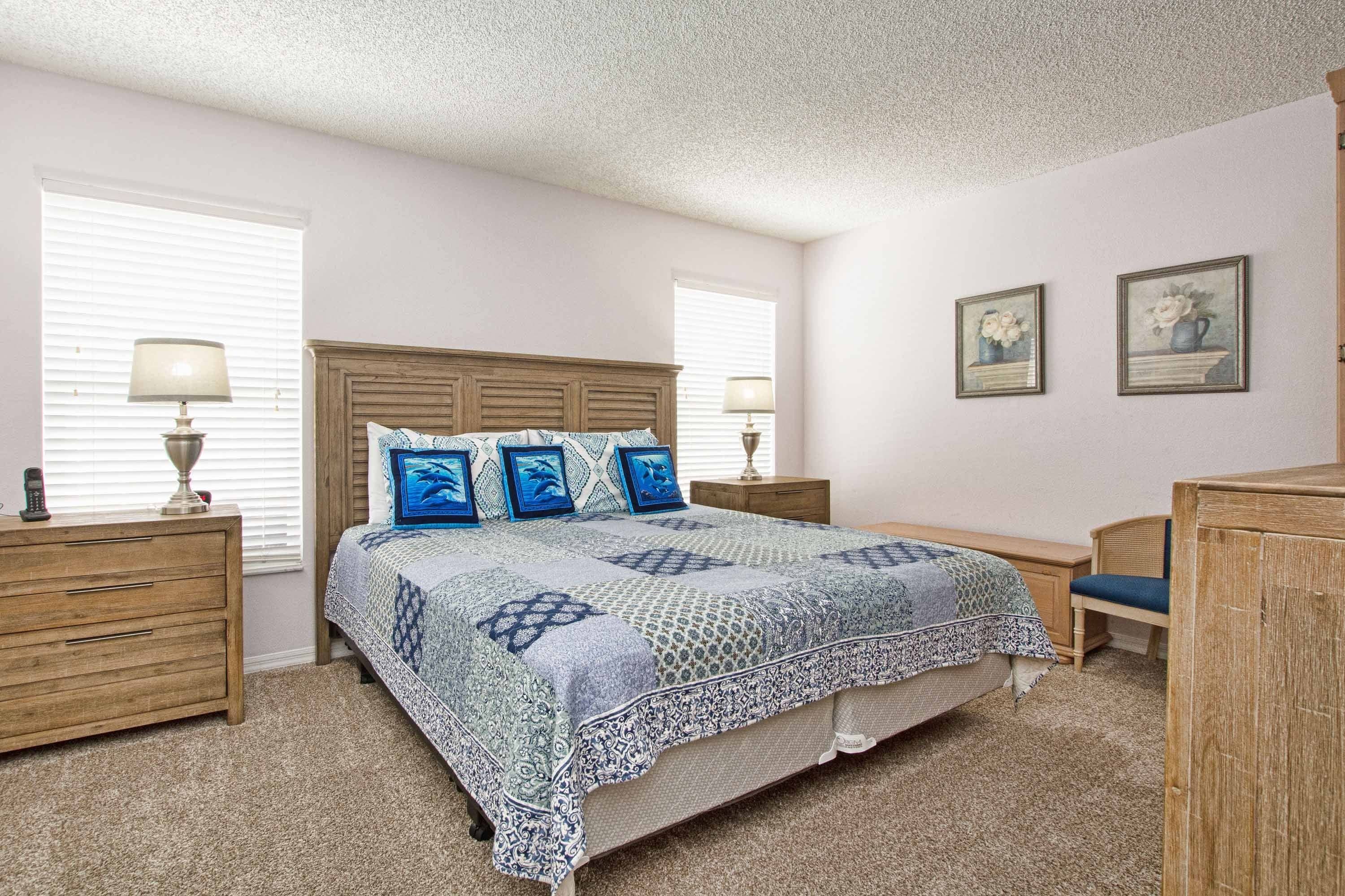 Une chambre à coucher avec une tête de lit en bois, une couvre-lit à motifs, deux lampes de chevet, une table de chevet en bois, une chaise et deux tableaux encadrés accrochés au mur.