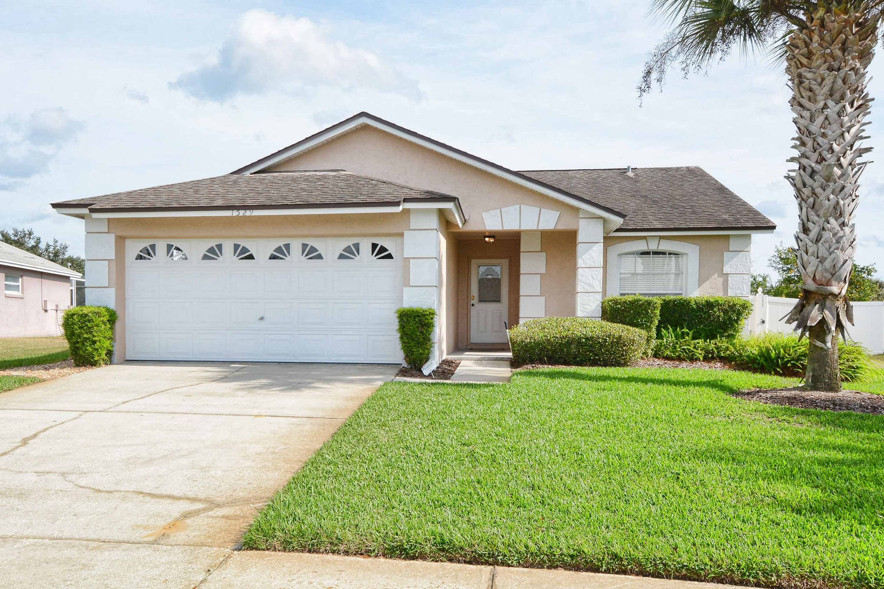 A single-story house with a garage, a front door, and a small front yard with a palm tree and shrubs.