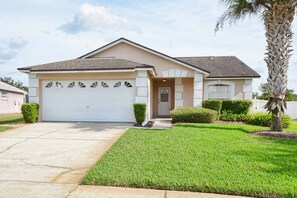 A single-story house with a garage, a front door, and a small front yard with a palm tree and shrubs.