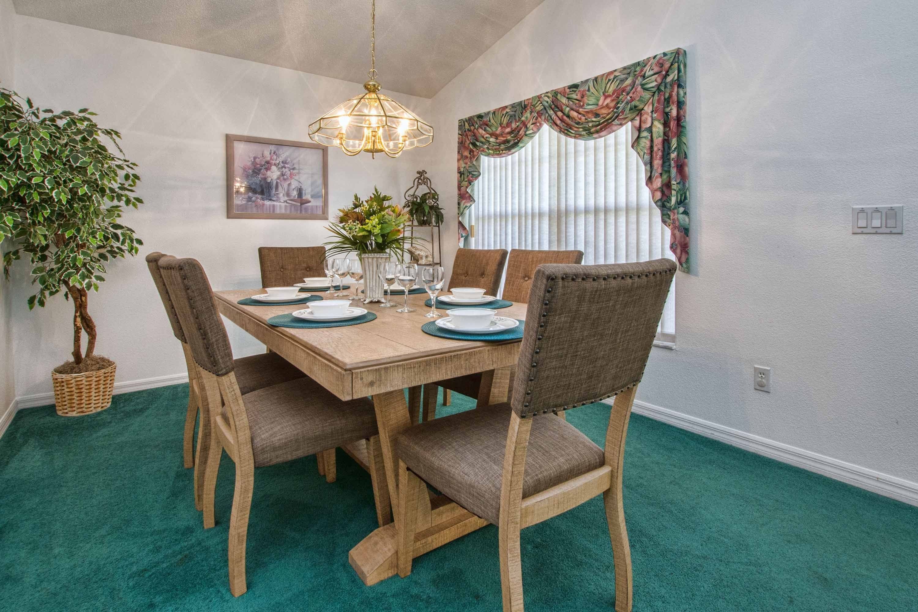 Une salle à manger avec une table en bois, six chaises, un lustre, une plante en pot et une fenêtre avec des rideaux.