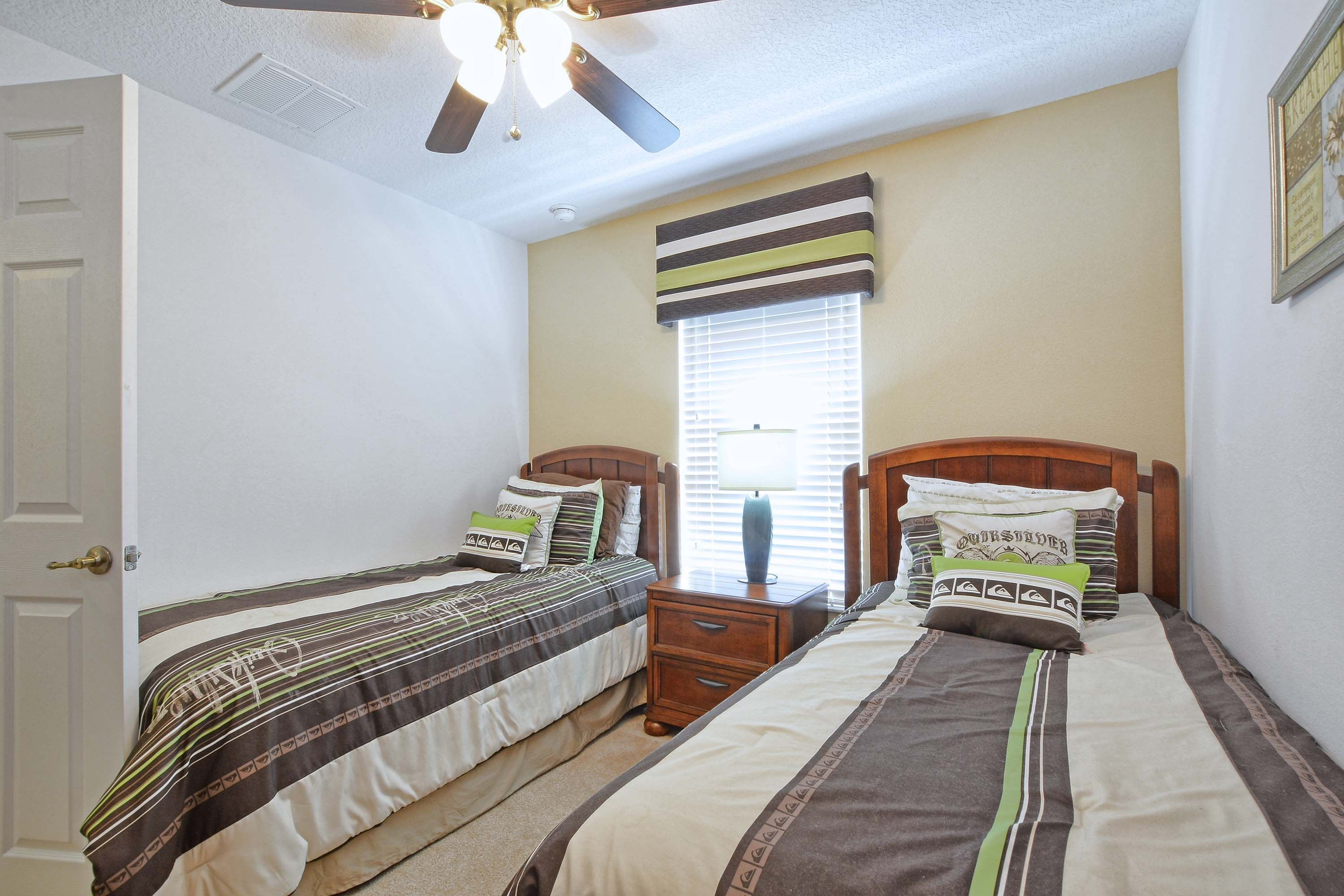 Une chambre avec deux lits simples, un ventilateur de plafond, une fenêtre avec des stores et une table de chevet avec une lampe.