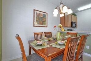 A dining room with a wooden table set for four, surrounded by chairs. There is a vase with flowers on the table, and a framed artwork hangs on the wall above.