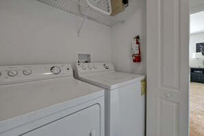 A laundry room with a front-loading washer and dryer, a shelf above the dryer, and a fire extinguisher mounted on the wall.