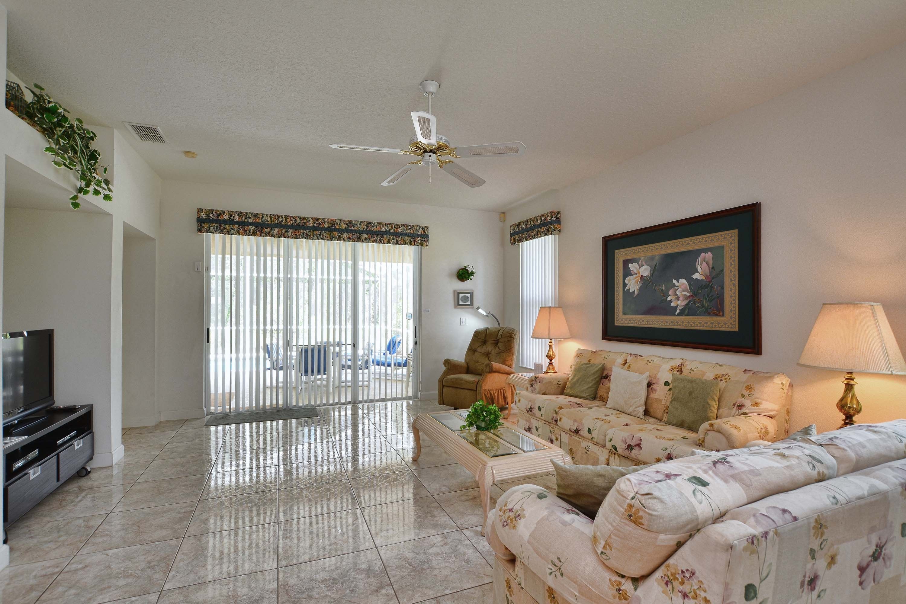 A living room with a sofa, a television, a ceiling fan, and a large window with blinds.