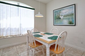 A dining area with a table, chairs, a painting, and a window with blinds.