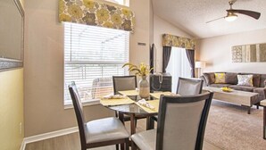 A dining area with a round table, chairs, and a vase with flowers. There is a window with blinds, a ceiling fan, and a couch in the background. - Emerald 4 bedrooms with Pool - 2201Em 4 Home by RedAwning (Kissimmee)
