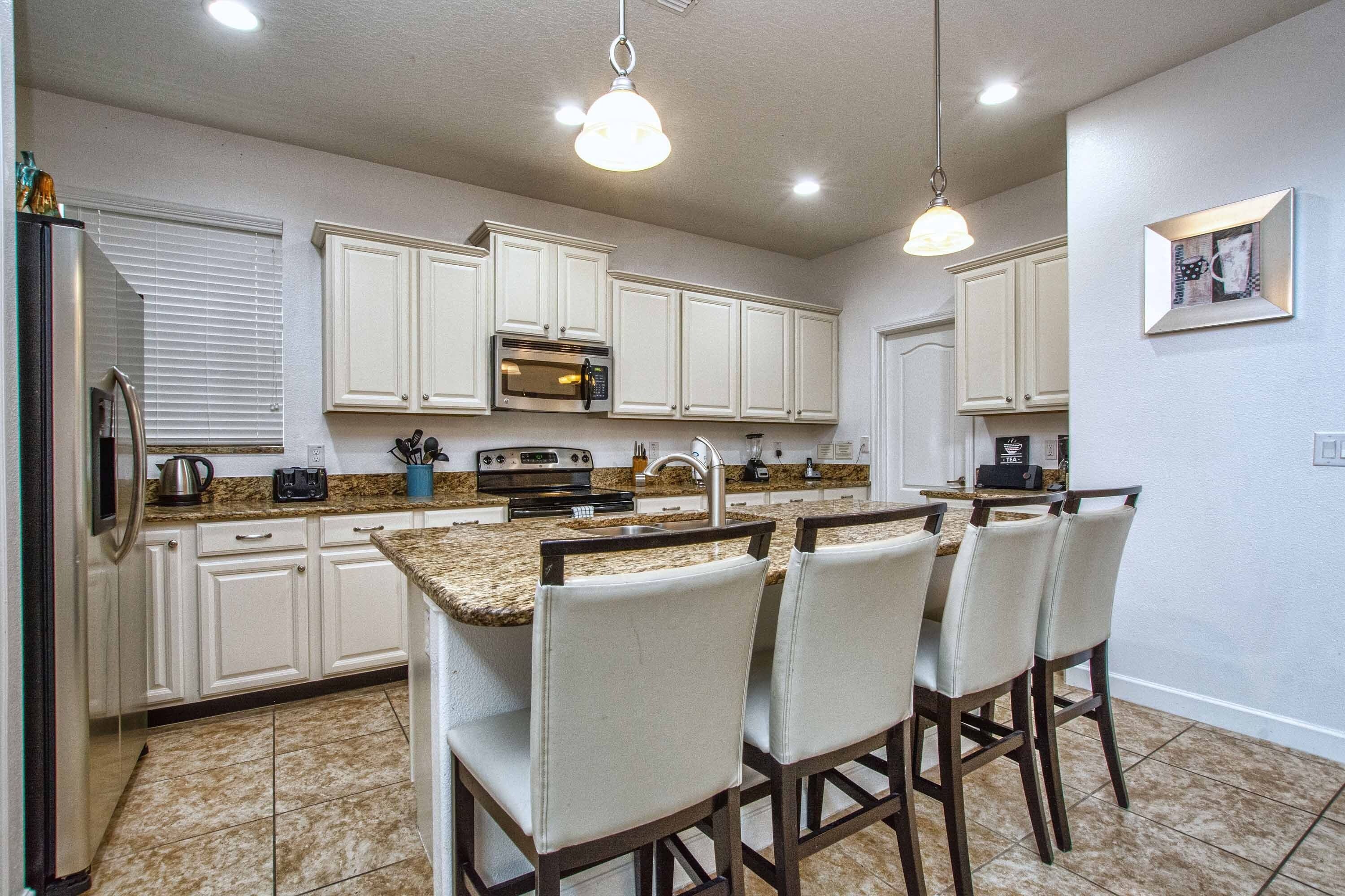 A kitchen with white cabinets, a built-in oven, a microwave, and a sink. There are four bar stools with cushioned seats.