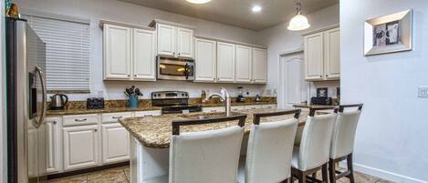 A kitchen with white cabinets, a built-in oven, a microwave, and a sink. There are four bar stools with cushioned seats.