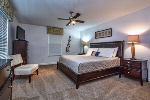 A bedroom with a bed, a desk, a chair, a ceiling fan, and a nightstand with a lamp.