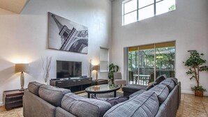 Ein modernes Wohnzimmer mit einem großzügigen Ecksofa, einem Flachbildfernseher, einem Couchtisch und einem großen Fenster mit Blick auf Grünflächen.