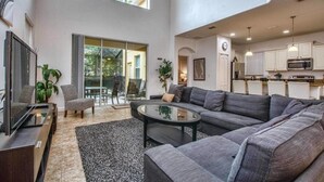Ein großzügiger Wohnbereich mit einer großen grauen Couch, einem Couchtisch und einem Fernseher. Eine Glastür führt auf eine Terrasse.