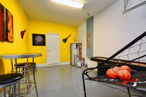 A room with a yellow wall, a dartboard, a guitar, a basketball hoop, and a table with chairs.