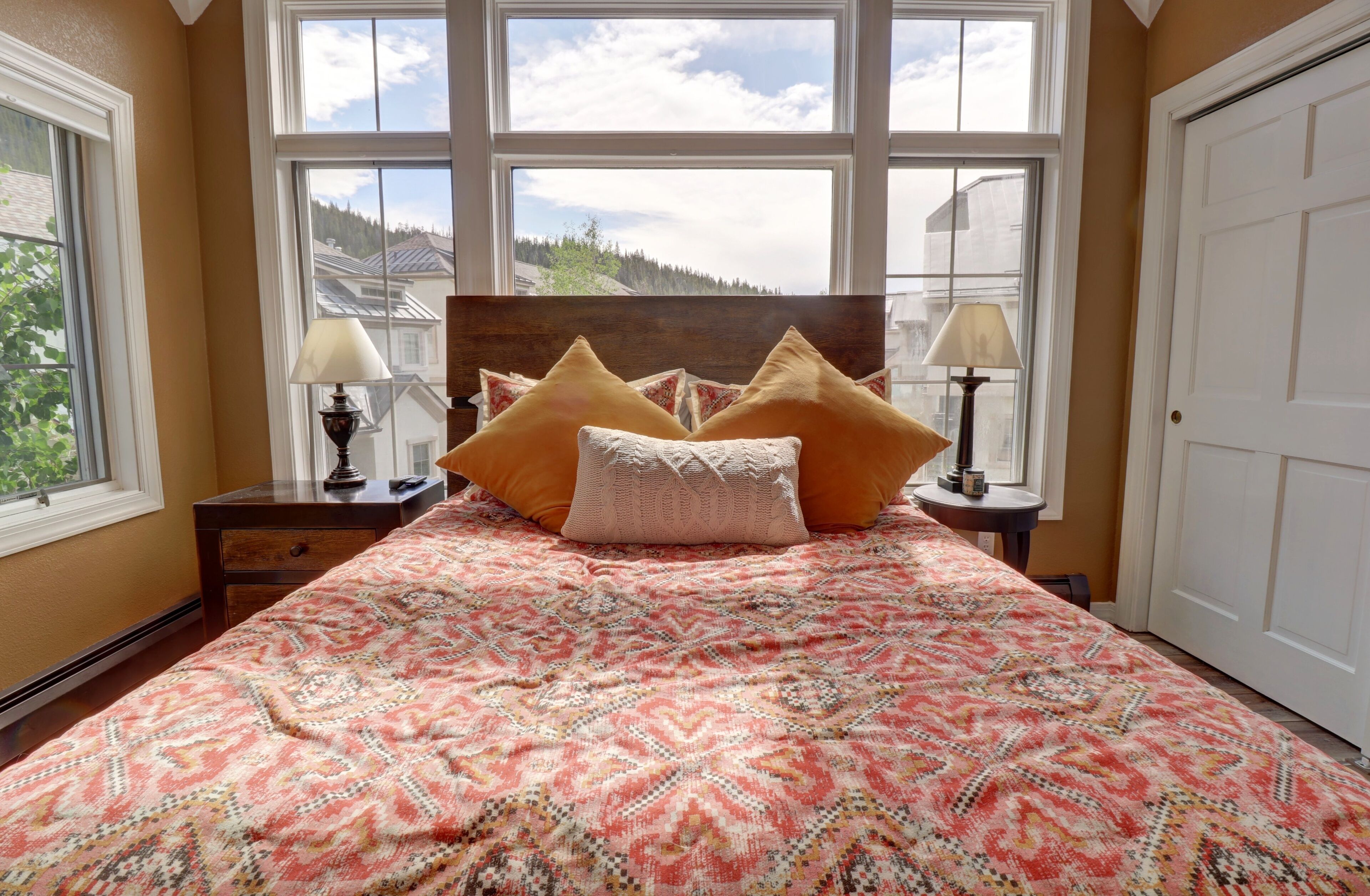 A bedroom with a large bed, two bedside tables, and a view of a forest through large windows.