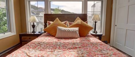 A bedroom with a large bed, two bedside tables, and a view of a forest through large windows.