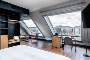 A modern hotel room with a large bed, a desk, and a chair. There are skylights and a view of the city.