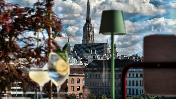 A table with a green lamp and two wine glasses, with a view of a historic building and a church in the background. - Radisson RED Hotel, Vienna (Vienna)
