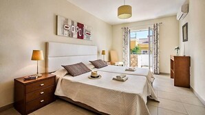 A bedroom with two beds, a nightstand, a lamp, a decorative wall piece, and a view of the outside.