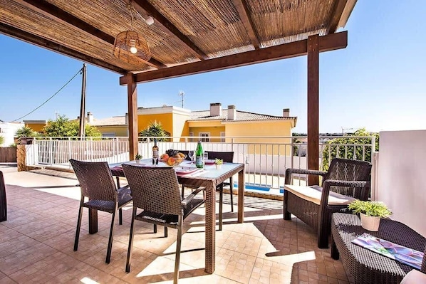 Une terrasse extérieure avec des meubles en osier, une table garnie de fruits et de boissons, et une vue sur les bâtiments en arrière-plan.