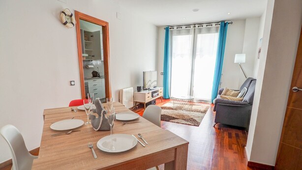 Un salon moderne avec un coin repas. La pièce est équipée d’une table à manger en bois, mise avec des assiettes, des couverts et des verres. On y trouve également un canapé et un fauteuil. La pièce est dotée d’une grande fenêtre avec des rideaux.