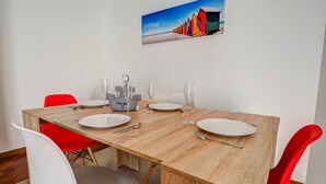 Un coin repas avec une table en bois, des chaises rouges et blanches, et un tableau représentant des cabanes de plage colorées accroché au mur.
