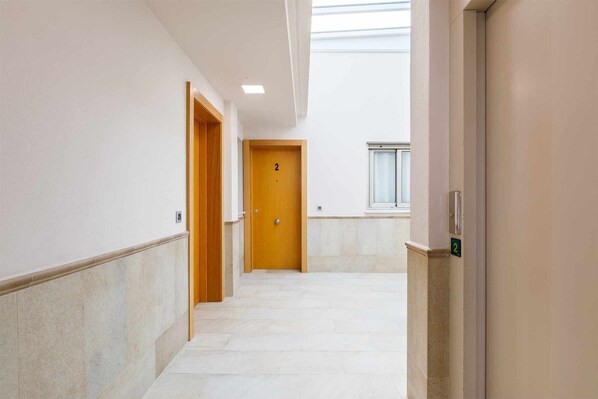 Un couloir avec une porte en bois portant le numéro « 2 », une fenêtre avec des stores et un sol carrelé.