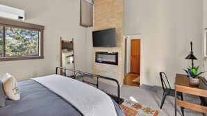 Une chambre avec un lit, un téléviseur fixé au mur en pierre, un bureau avec une lampe et une fenêtre donnant sur les arbres.