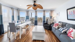 A modern living room with a dining area, a ceiling fan, and a large window with curtains. - Silver Sands 102S Direct Ocean Front, Just steps to the Beach! (Ocean City)