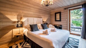 A wooden-paneled bedroom with a large bed, a chandelier, a side table with a lamp, and a view through a glass door to an outdoor area. - PADOUK- Apartment w/ hot tub in Courchevel Moriond (Courchevel)