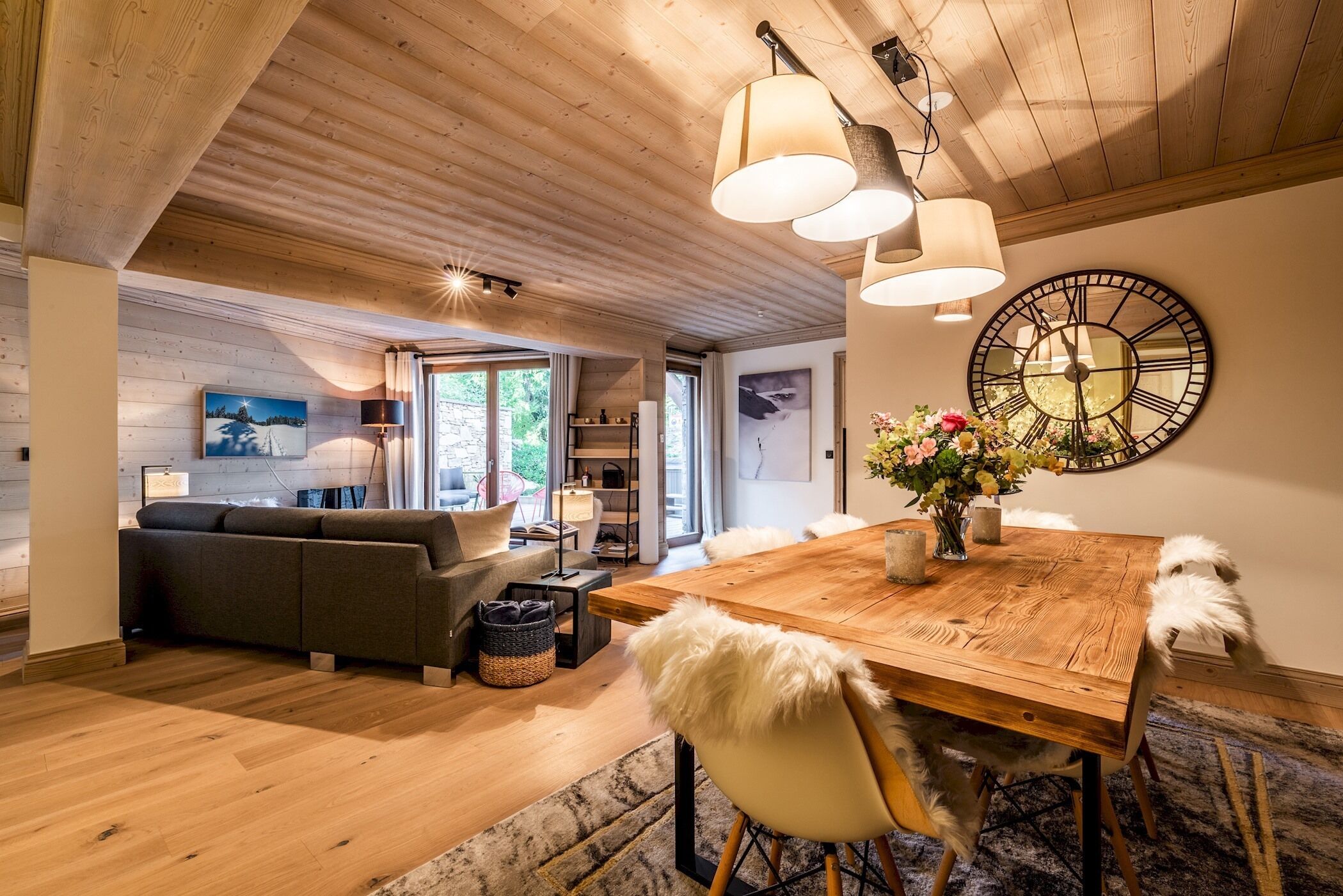 A spacious living area with a wooden ceiling, a dining table with chairs, a sofa, and a large wall clock.
