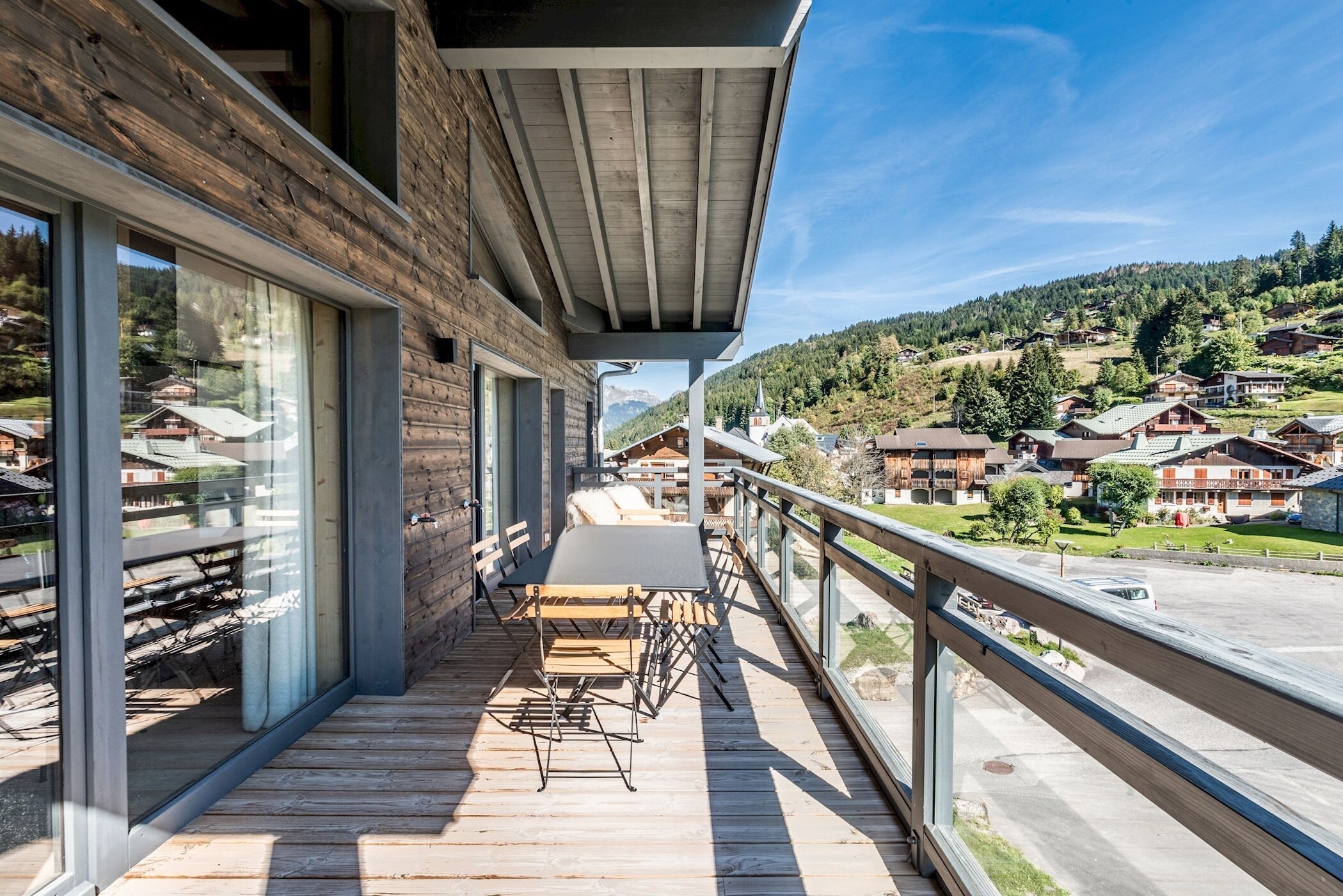 Una terraza de madera con una mesa y sillas, con vista a un barrio residencial con casas y una calle.
