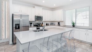 A modern kitchen with white cabinets, stainless steel appliances, and a large island with clear bar stools. - Solara Resort 4 Bedroom Townhome- Private Pool (Kissimmee)