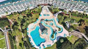 An aerial view of a hotel with a central swimming pool, multiple balconies, and surrounding greenery.