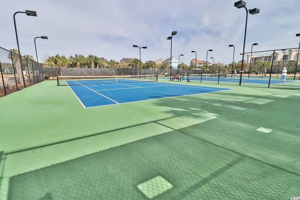 An outdoor tennis court with multiple courts, surrounded by a fence, and equipped with overhead lighting.