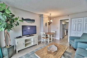 A living room with a television, a dining table, chairs, a coffee table, a potted plant, and a door leading to the kitchen.