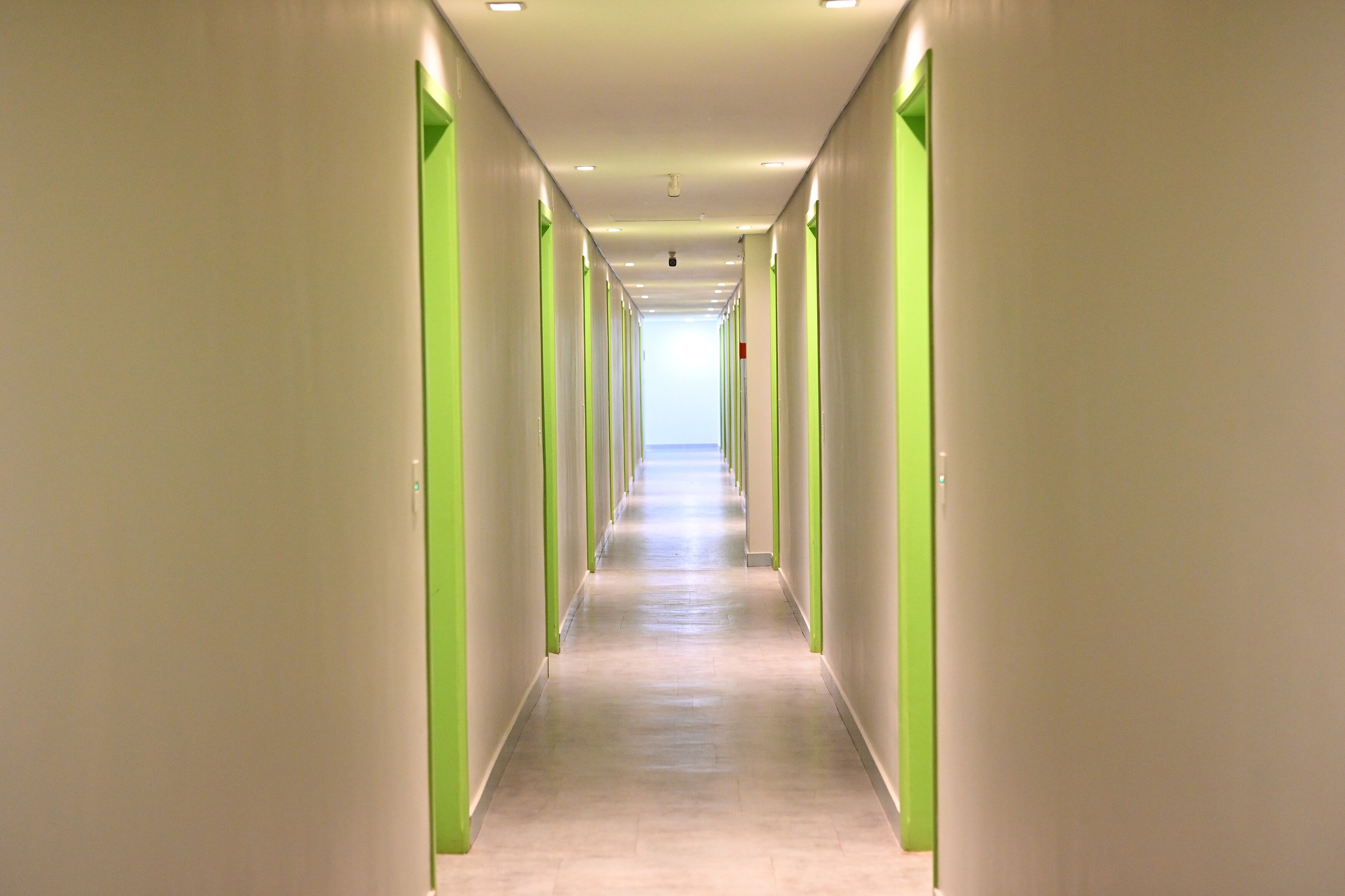 A long, narrow hallway with green doors on both sides and a light-colored floor.